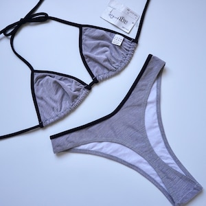 Könnte beinhalten: Graues Bikini-Set mit schwarzer Paspelierung. Das Oberteil ist im Dreiecks-Stil mit dünnen Trägern gehalten, und das Unterteil ist klassisch geschnitten. Der Stoff hat eine dezente Streifenstruktur. Ein Etikett ist am Oberteil befestigt.