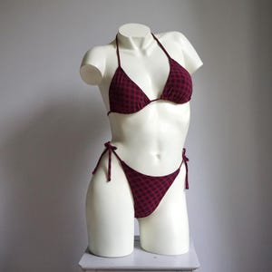 Peut inclure: Ensemble de bikini deux pièces à carreaux bordeaux et noirs. Le haut est de style triangle avec des bretelles, et le bas a des liens sur les côtés. Présenté sur un mannequin blanc.