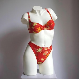 May include: A red bikini set with a floral pattern, displayed on a white mannequin. The two-piece swimsuit features a bra top with adjustable straps and a high-cut bottom. The floral design includes yellow, orange, and green accents.