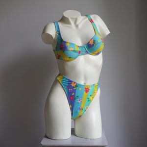 Pode incluir: Um conjunto de biquíni de duas peças com padrão floral. A parte de cima é estilo balconet com alças ajustáveis. A parte de baixo é de corte alto. O tecido apresenta riscas verticais em azul, amarelo e roxo, adornadas com flores coloridas.