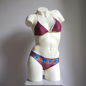 Op de afbeelding: Een tweedelige bikiniset op een mannequin. De top is een driehoekmodel met een donkerrode basis en witte stippen, afgewerkt met wit. De broek heeft een bloemenpatroon met blauwe en oranje bloemen op een donkerrode achtergrond.