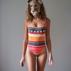 Op de afbeelding: Een vrouw draagt een gestreept badpak met een leeuwenmasker. Het badpak heeft een rood, oranje, bruin en zwart gestreept patroon met witte accenten.
