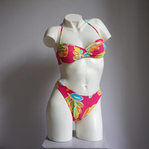 May include: A two-piece bikini set with a vibrant floral pattern. The halter-style top and high-cut bottoms feature a hot pink base adorned with colorful leaf-like designs in shades of orange, yellow, blue, and green. The bikini is displayed on a white mannequin.