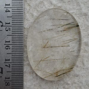 Puede incluir: Gema de cuarzo transparente de forma ovalada con inclusiones negras y doradas. La gema está junto a una regla metálica. El cuarzo mide aproximadamente 4,4 cm de largo.