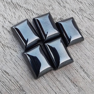 May include: Five polished black gemstone cabochons arranged in a diamond shape. Each stone has a rectangular shape with beveled edges, reflecting light. The gemstones are set against a weathered, grey wooden surface.