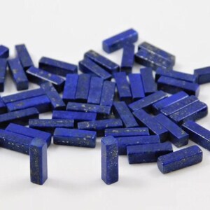 Puede incluir: Una colección de pequeñas piedras preciosas rectangulares de lapislázuli. Las piedras son de color azul intenso con motas doradas y están dispersas sobre una superficie blanca. Las piedras preciosas están cortadas en forma rectangular.