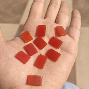 May include: A hand holding ten rectangular, polished, translucent, red-orange gemstones. The stones are evenly distributed across the palm, showcasing their smooth, glossy surfaces. The background is a neutral, checkered pattern.