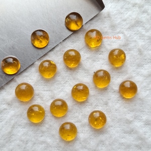 Peut inclure: Une collection de pierres précieuses rondes de couleur ambre. Les gemmes sont lisses et polies, avec une qualité translucide. Elles sont dispersées sur une surface blanche, avec une surface métallique en arrière-plan. Les gemmes varient légèrement en couleur.