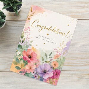 Puede incluir: Una tarjeta de felicitación rectangular con un diseño floral de acuarela en tonos rosa, morado y verde, con el texto dorado "Congratulations! SO HAPPY FOR YOU AND ALL THAT YOU ACHIEVE" impreso en el centro, acompañada de dos pequeñas plantas suculentas.