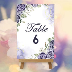 May include: A white table number card with the text "Table 6" in elegant script and a bold font. The card is decorated with watercolor-style purple roses and greenery, and is displayed on a small wooden easel.