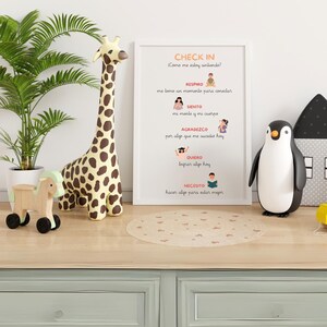 Puede incluir: Un póster enmarcado blanco con el título "Check In" y una lista de preguntas en español: "Como me estoy sintiendo?", "Respiro", "Siento", "Agradezco", "Quiero", "Necesito". El póster está en un estante de madera con un juguete de jirafa y un juguete de pingüino.