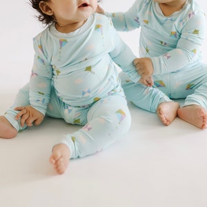 K&ouml;nnte beinhalten: Zwei Babys tragen passende hellblaue Pyjamas mit einem bunten Drachenmuster. Die Pyjamas haben lange &Auml;rmel und lange Hosen.