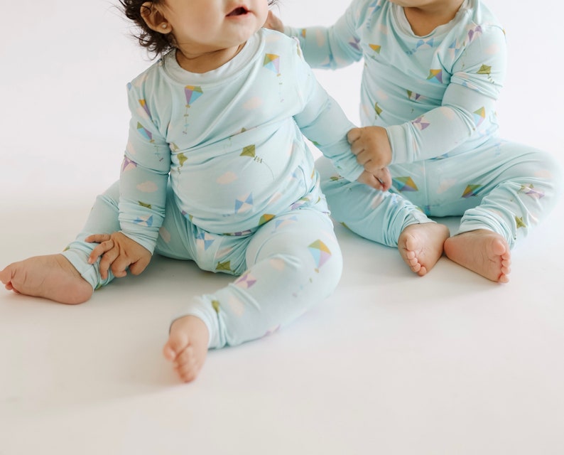 K&ouml;nnte beinhalten: Zwei Babys tragen passende hellblaue Pyjamas mit einem bunten Drachenmuster. Die Pyjamas haben lange &Auml;rmel und lange Hosen.