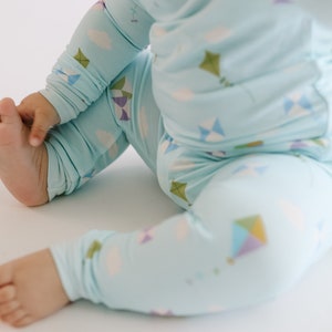 K&ouml;nnte beinhalten: Ein Baby tr&auml;gt einen hellblauen Schlafanzug mit einem Drachenmuster. Der Schlafanzug hat lange &Auml;rmel und lange Beine. Das Baby sitzt auf einer wei&szlig;en Oberfl&auml;che.