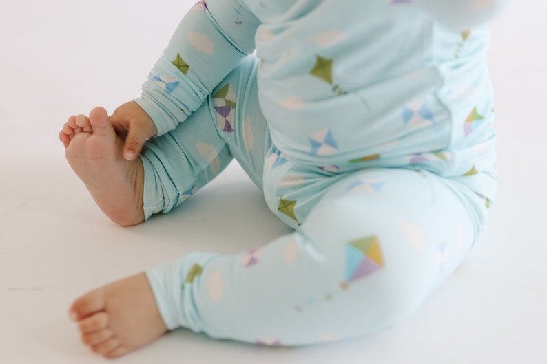 K&ouml;nnte beinhalten: Ein Baby tr&auml;gt einen hellblauen Schlafanzug mit einem Drachenmuster. Der Schlafanzug hat lange &Auml;rmel und lange Beine. Das Baby sitzt auf einer wei&szlig;en Oberfl&auml;che.