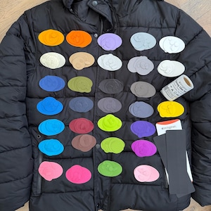 Könnte beinhalten: Eine schwarze, gesteppte Daunenjacke mit einer Sammlung zahlreicher ovaler Klebeflicken in verschiedenen Unifarben wie Orange, Blau, Grün, Pink und Lila, ergänzt durch eine Rolle Zengear-Klebeband und zwei schwarze, rechteckige Streifen.
