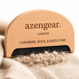 May include: A semi-circular wooden fabric comb with a black mesh edge, featuring the engraved text "azengear. LONDON CASHMERE, WOOL & KNITS COMB". The tool is positioned on a beige knitted garment, with a small pile of removed fabric pills below it.
