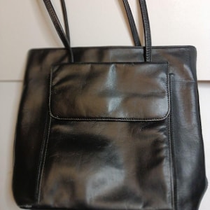 Könnte beinhalten: Eine schwarze Ledertasche mit zwei langen Henkeln und einer Vordertasche. Die Tasche hat eine rechteckige Form und eine glatte, glänzende Oberfläche. Die Henkel sind dünn und scheinen aus dem gleichen Material wie die Tasche zu sein.