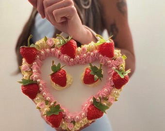 May include: Heart-shaped handbag with a strawberry and cream design. The bag features a white center, pink and yellow frosting, and is adorned with red strawberries and pearl-like beads. A unique, whimsical accessory.