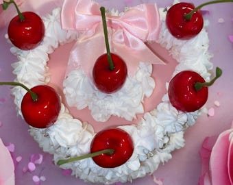 Puede incluir: Un pastel rosa y blanco decorado con cerezas rojas y un lazo de satén rosa. El pastel está rodeado de confeti rosa.