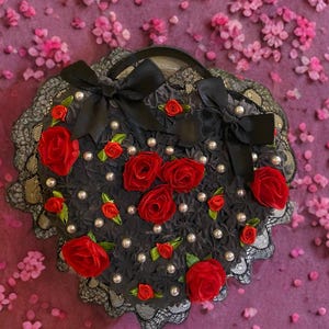 May include: A black, scalloped-edge handbag with red roses, pearls, and black bows. The bag has a lace trim and a handle. The background is a purple surface with pink flower petals.