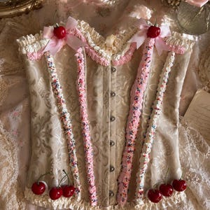May include: A beige corset with a damask pattern, decorated with faux whipped cream piping, colorful sprinkles, and red cherry accents. The garment features pink ribbon bows at the top, a ruffled hem, and a front closure with silver metal buttons.