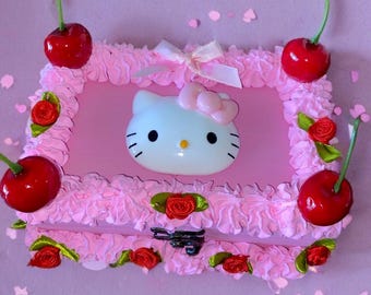 Puede incluir: Un marco rectangular rosa con una cara de Hello Kitty en el centro, rodeado de un borde rosa en forma de glaseado. Cerezas rojas con tallos verdes se colocan en cada esquina. Pequeñas rosas rojas y un lazo blanco completan el diseño decorativo.