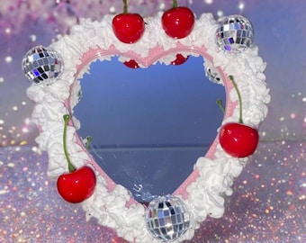 Puede incluir: Un espejo rosa en forma de corazón con un borde blanco, esponjoso, similar a la crema batida. Hay tres cerezas rojas y cuatro acentos de bola de discoteca plateada alrededor del espejo.