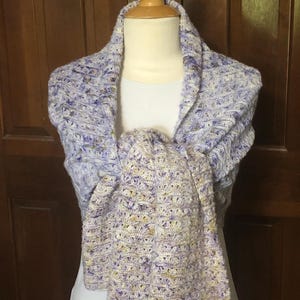 May include: A purple and white crocheted shawl with a textured pattern. The shawl is draped over a white mannequin.