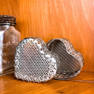 May include: Two open, heart-shaped, silver-colored tea infusers with a decorative filigree design. The infusers are made of a perforated metal. A glass jar with a metal lid is in the background, all on a wooden surface.