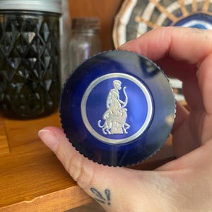 May include: A dark blue lid with a silver emblem featuring a figure with a bow and arrow, and a lion. The lid is round and has a ridged edge. The background includes a glass jar and other containers.