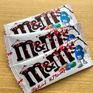 May include: Three individually wrapped chocolate candy bars with the brand name "M&M's" printed in black on a white background. The candy bars are decorated with pink cherry blossoms and feature a blue M&M character on the front. The bottom candy bar has the text "Merci Richey" printed in red.