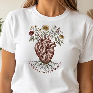 May include: A white t-shirt featuring a graphic of an anatomical heart with roots at the bottom and flowers blooming from the top. The text "HOPE TAKES ROOT" is printed across the heart, with "CHILDHOOD CANCER AWARENESS" curved underneath.