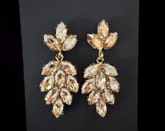 May include: A pair of gold-toned drop earrings with a leaf-like design. Each earring features a cluster of champagne-colored, marquise-cut crystals. The earrings are displayed on a black background.