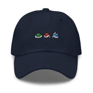 Op de afbeelding: Een marineblauwe baseballcap met geborduurde pixelontwerpen van een groene schildpad, een rode paddenstoel en een blauw kasteel. De cap heeft een gebogen klep en een knoop aan de bovenkant.
