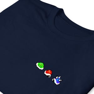 Può includere: Una t-shirt blu navy con un design pixelato di tre power-up di Super Mario Bros. di diversi colori. I power-up sono verde, rosso e blu.