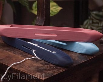 May include: Three colorful weaving shuttles in pink, blue, and navy blue rest on a wooden surface. The shuttles are smooth and oblong, with a slot for yarn. A white yarn is threaded through the navy blue shuttle.