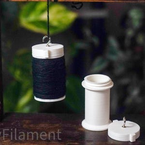 Puede incluir: Una bobina de hilo negro suspendida de un gancho, con tapa y base blancas. Junto a ella, un recipiente cilíndrico blanco con tapa a juego. Los artículos están sobre una superficie de madera, con un fondo verde borroso.