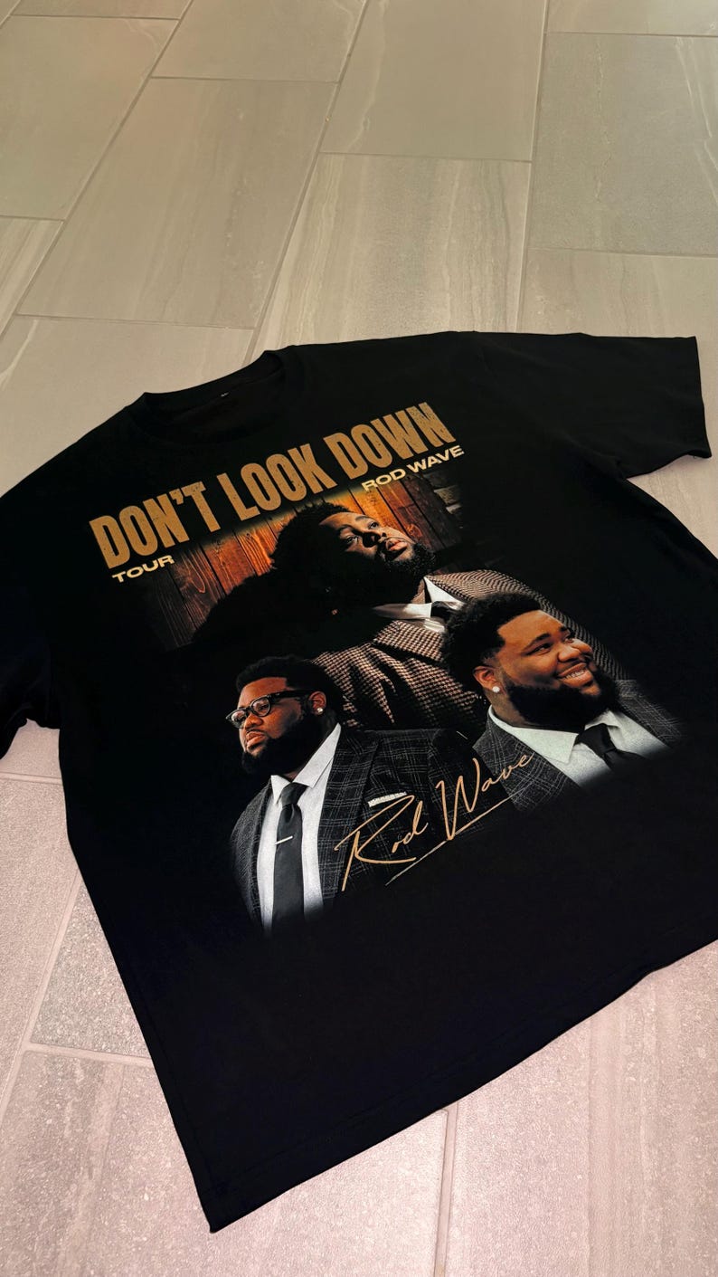 May include: A black short-sleeve t-shirt featuring a graphic print of Rod Wave in a plaid suit. The text reads DON'T LOOK DOWN TOUR and ROD WAVE in gold lettering. A signature in gold script is positioned over the lower right portion of the graphic.