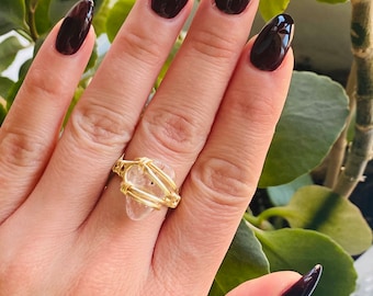 May include: A gold wire wrapped ring featuring a clear quartz stone. The ring is worn on a hand with dark burgundy nail polish.