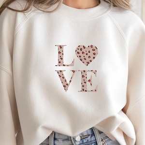Op de afbeelding: Crèmekleurige sweatshirt met het woord "LOVE" in een roze luipaardprint. Het hart in het woord "LOVE" is ook in de luipaardprint. De sweatshirt heeft lange mouwen en een ronde hals.