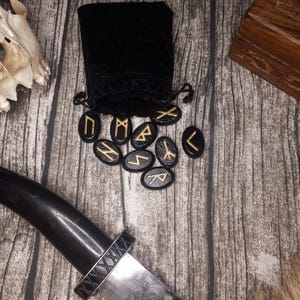 Może przedstawiać: Czarna aksamitna torebka z zestawem czarnych kamieni runicznych z złotymi napisami. Runy są rozłożone na drewnianej powierzchni.