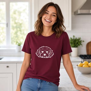 Puede incluir: Una camiseta de manga corta en color beige claro con una ilustración de línea negra centrada de una cara de perro esponjoso con orejas caídas y un hocico detallado. La prenda tiene un cuello redondo clásico y un dobladillo recto.