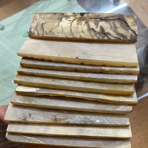 May include: A stack of eight thin, rectangular pieces of light brown, white, and gray wood. The wood has a natural, organic pattern.