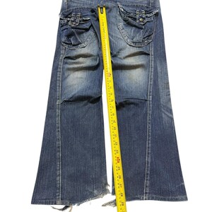May include: Blue denim jeans with flared legs and distressed detailing. The jeans feature two large pockets on the back with button closures. A yellow measuring tape is placed along the length of the jeans, showing measurements in inches.