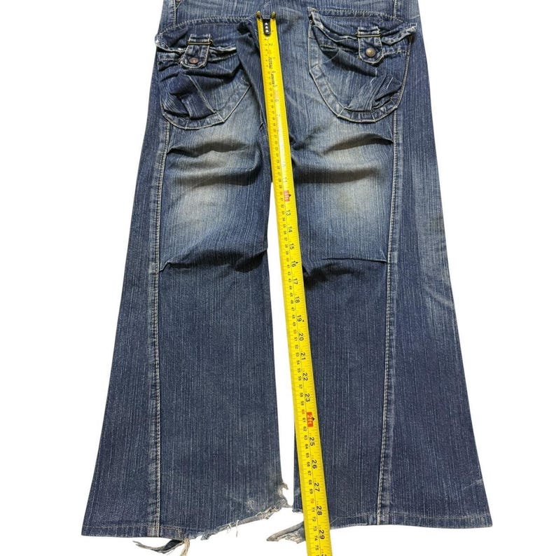 May include: Blue denim jeans with flared legs and distressed detailing. The jeans feature two large pockets on the back with button closures. A yellow measuring tape is placed along the length of the jeans, showing measurements in inches.