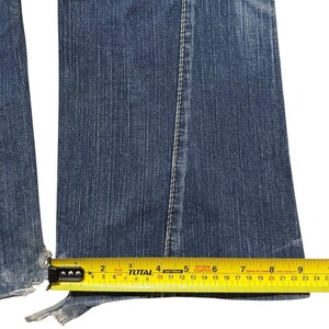 May include: Close-up of blue denim jeans with a frayed hem. A yellow measuring tape shows the width of the jeans at the hem is approximately 9 inches. The jeans have a white seam down the center.