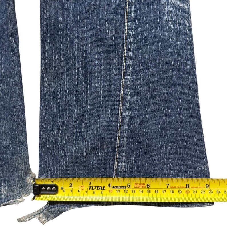 May include: Close-up of blue denim jeans with a frayed hem. A yellow measuring tape shows the width of the jeans at the hem is approximately 9 inches. The jeans have a white seam down the center.