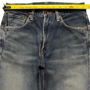 May include: Blue denim jeans with a faded wash, featuring a button fly, copper-toned rivets, and five pockets. A yellow measuring tape is positioned across the waistband, indicating a width of approximately 15 inches from edge to edge.