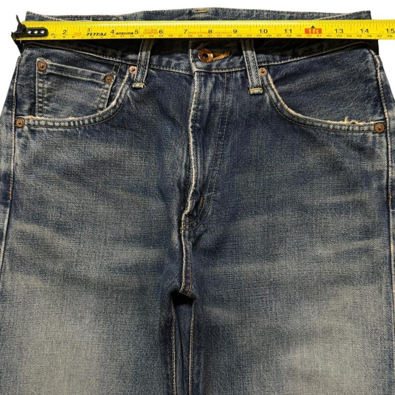 May include: Blue denim jeans with a faded wash, featuring a button fly, copper-toned rivets, and five pockets. A yellow measuring tape is positioned across the waistband, indicating a width of approximately 15 inches from edge to edge.